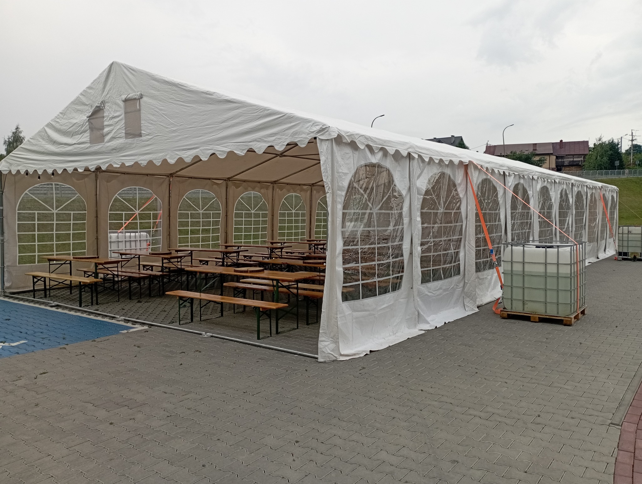 Rozłożony biały namiot z oknami, wewnątrz drewniane stoły i ławki. Na zewnątrz zbiorniki z wodą. Widok na szarą kostkę brukową i budynki w tle.