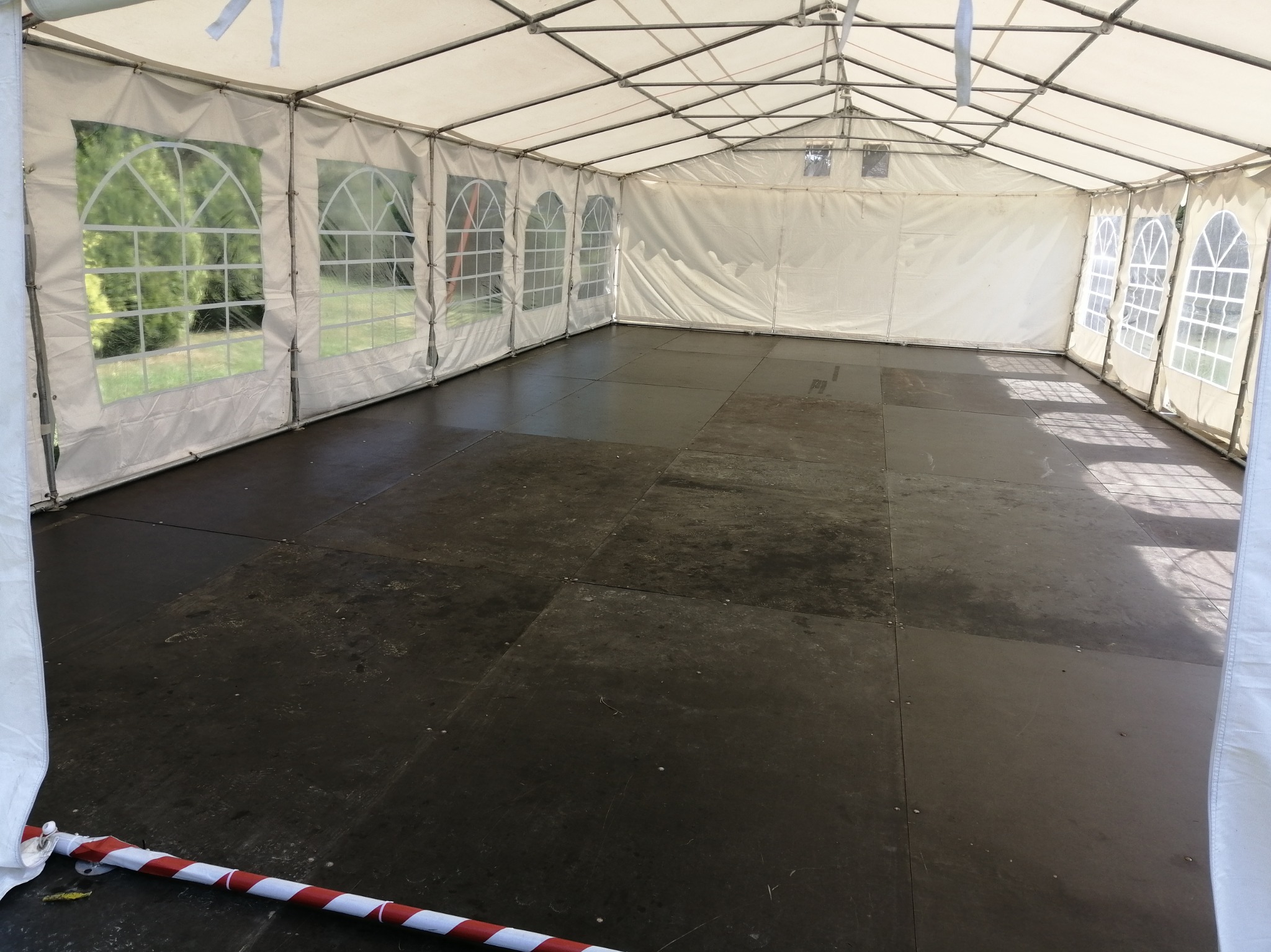 Wnętrze białego namiotu eventowego z podłogą z ciemnych płyt i oknami z widokiem na zieleń. Czerwono-biała taśma zabezpieczająca przy krawędzi.