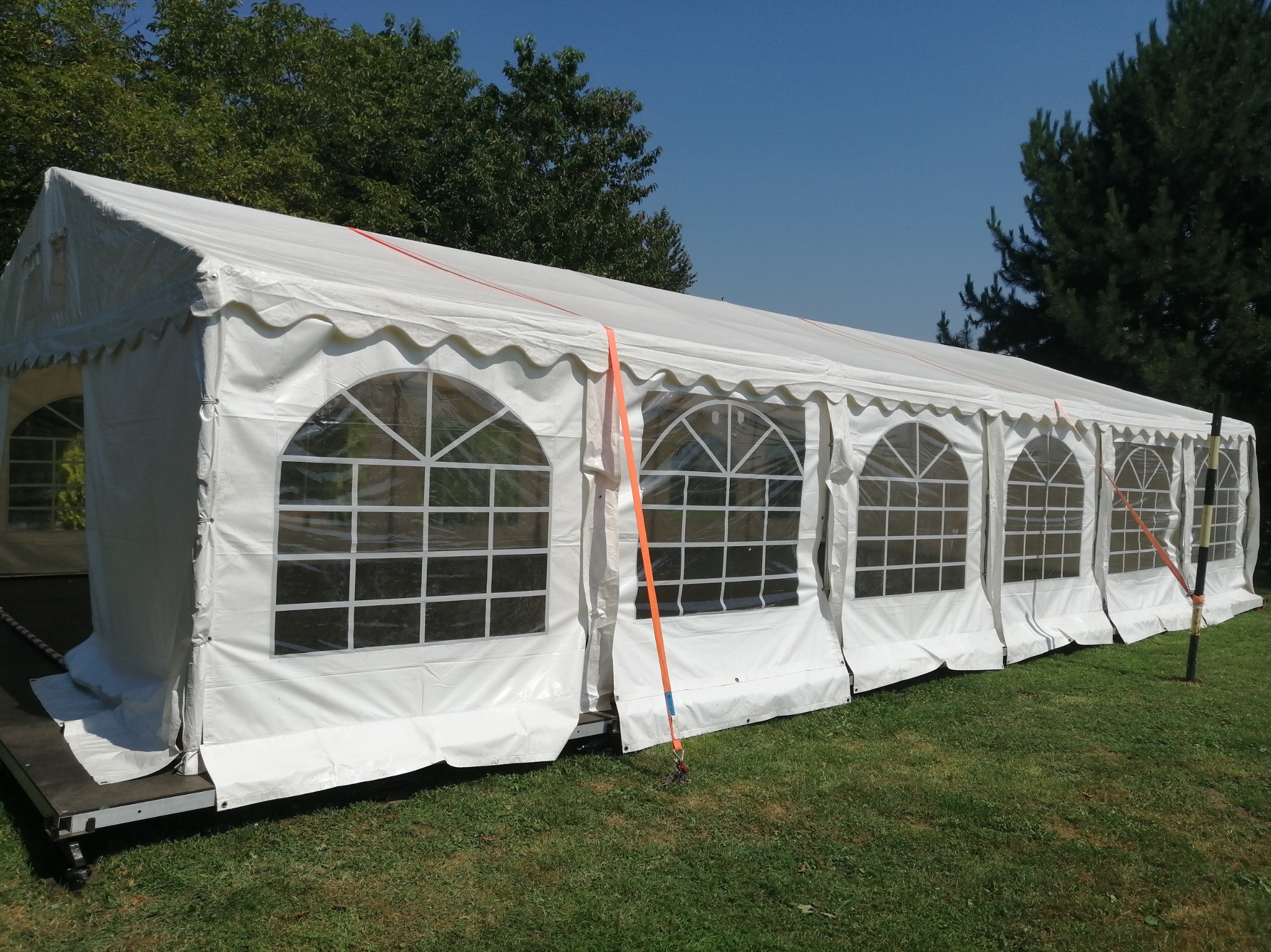 Rozłożony biały namiot eventowy z oknami na trawie, zabezpieczony pomarańczowymi pasami, w tle drzewa i błękitne niebo.