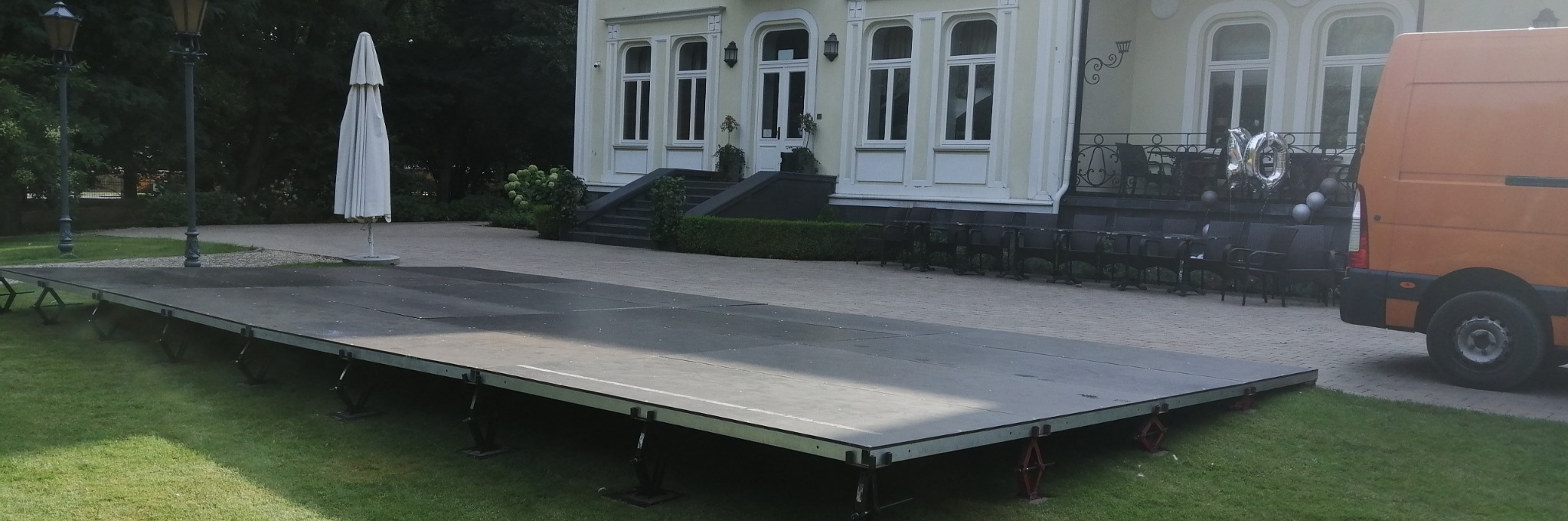 Rozłożona scena plenerowa na trawie przed budynkiem z okazji 10-lecia, obok zaparkowany pomarańczowy bus. Krzesła i balony w tle sugerują przygotowania do wydarzenia.