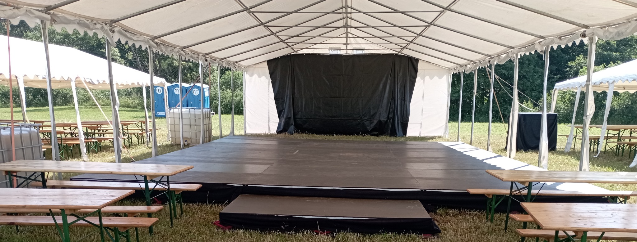 Scena pod namiotem z czarnym tłem, stoły piknikowe i toalety przenośne w tle. Przygotowania do imprezy plenerowej.