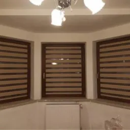 Trzy brązowe okna z poziomymi żaluzjami w odcieniach beżu i brązu, widoczne w jasnym pokoju z białymi ścianami i kaloryferem.