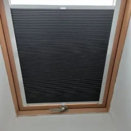 Ciemnoszara, plisowana roleta wewnętrzna zamontowana w oknie dachowym z drewnianą ramą, widoczny mechanizm sterujący.