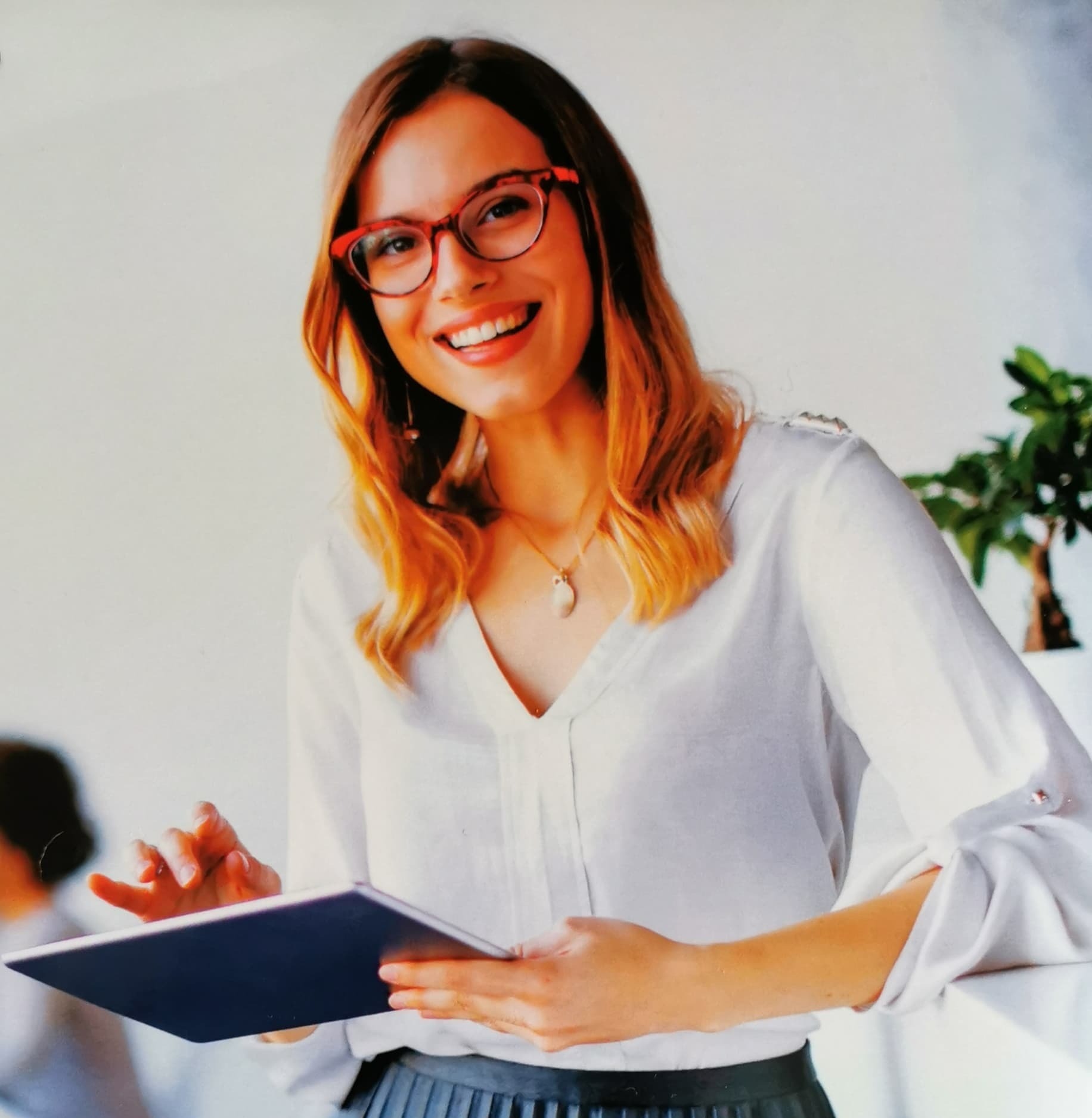 Uśmiechnięta kobieta w okularach z czerwoną oprawką, w białej bluzce, trzymająca tablet i opierająca się o białą ścianę, z rośliną doniczkową w tle.