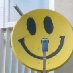 Żółta antena satelitarna pomalowana na kształt uśmiechniętej buźki, zamontowana na tle białego balkonu i okna.