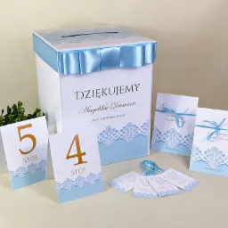 Biała skrzynka na koperty z niebieską wstążką i napisem 'Dziękujemy Angelika & Damian' otoczona zaproszeniami ślubnymi i numerami stołów w kolorystyce błękitu i złota.
