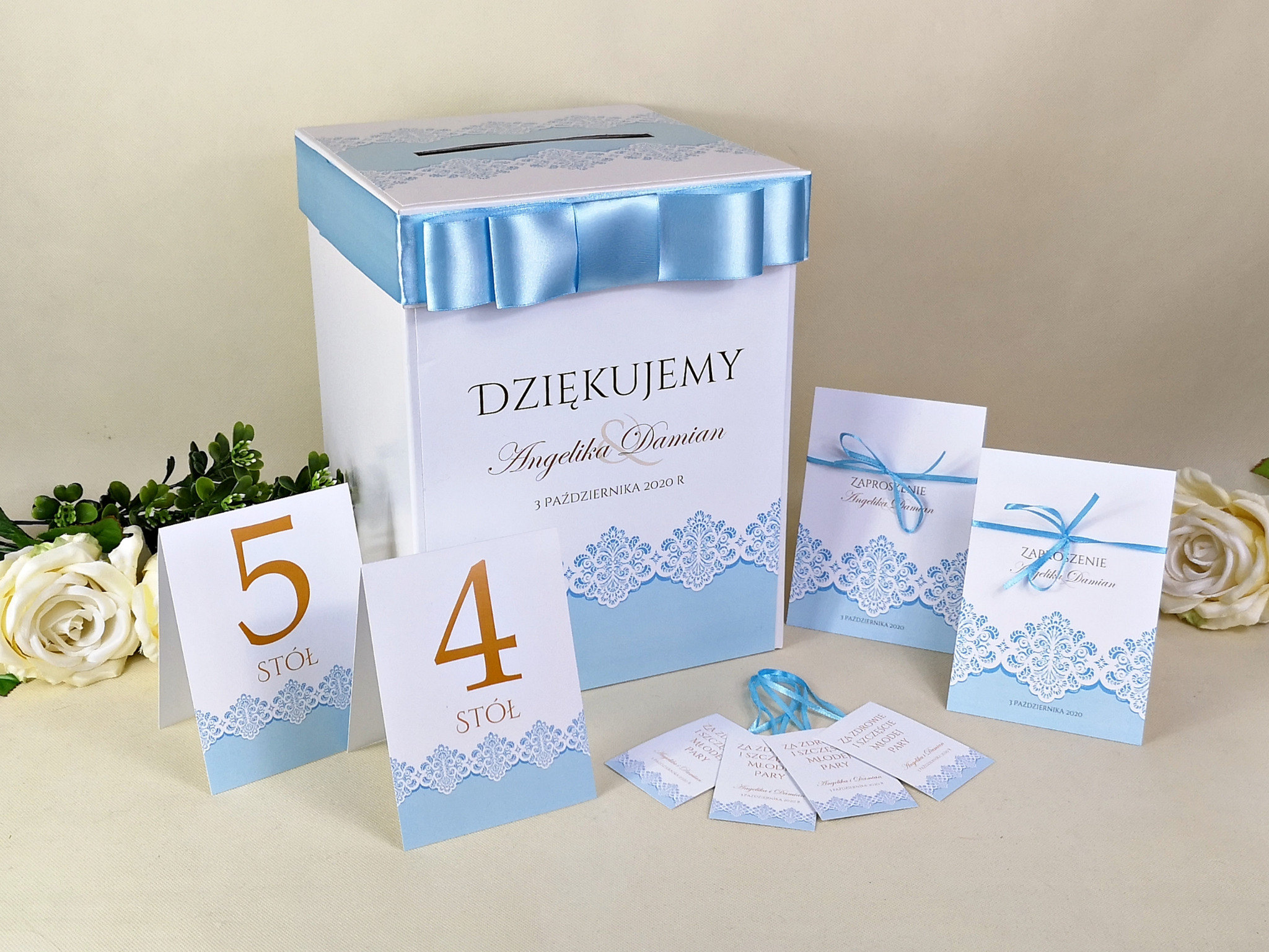 Biała skrzynka na koperty z niebieską wstążką i napisem 'Dziękujemy Angelika & Damian' otoczona zaproszeniami ślubnymi i numerami stołów w kolorystyce błękitu i złota.