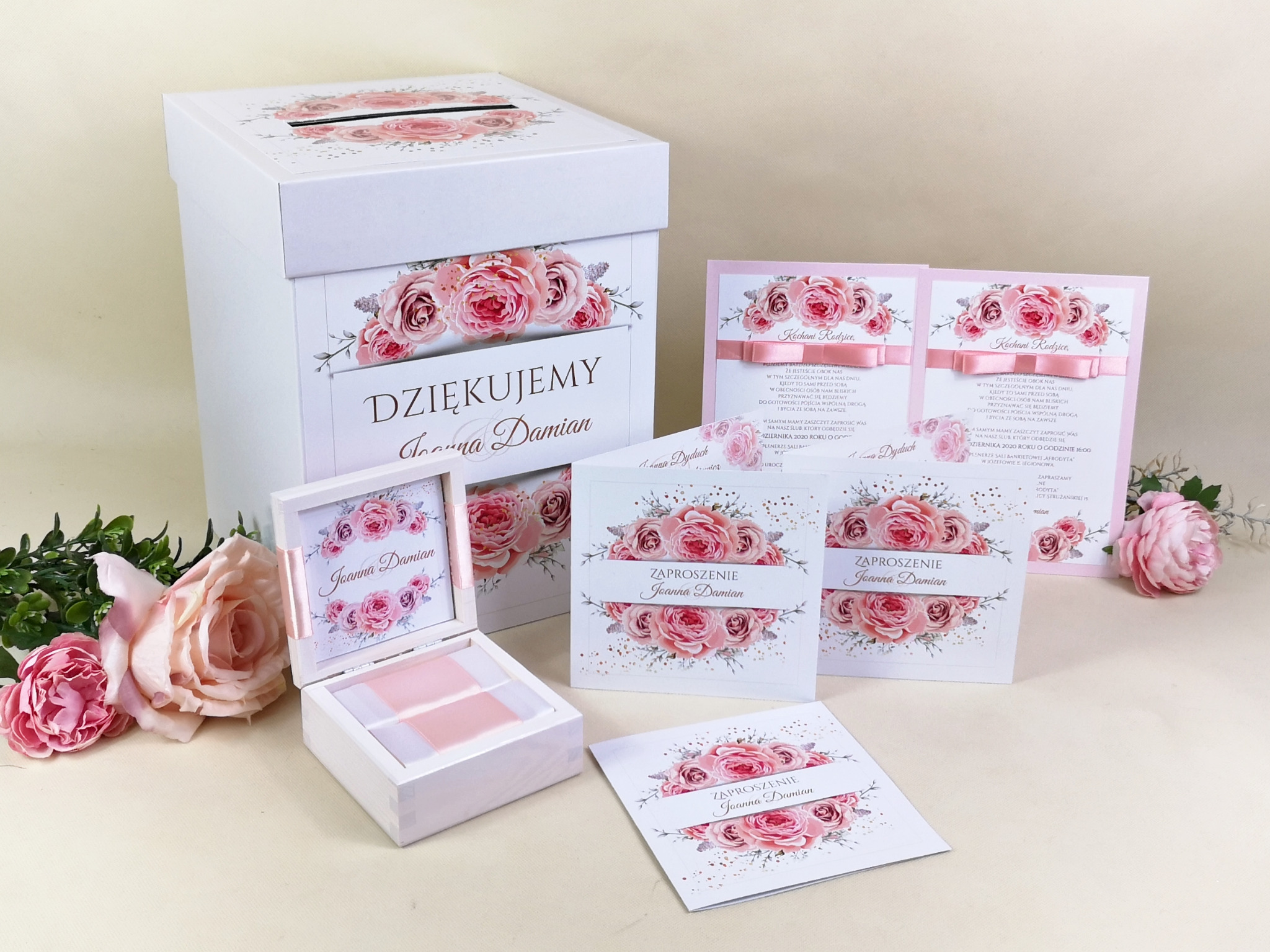 Białe, kwadratowe pudełko na koperty z napisem 'Dziękujemy Joanna i Damian' oraz zaproszenia ślubne z motywem różowych róż, całość w jasnej tonacji.