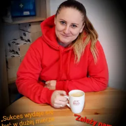 Kobieta w czerwonej bluzie z kapturem siedzi przy stole z kubkiem kawy z logo Generali, w tle półka z apteczką pierwszej pomocy i stosem pudełek, na stole cytat motywacyjny