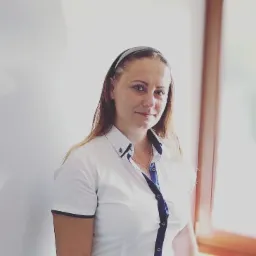 Uśmiechnięta kobieta w białej koszuli z granatowymi akcentami i opaską na włosach, stojąca na tle jasnej ściany i okna.