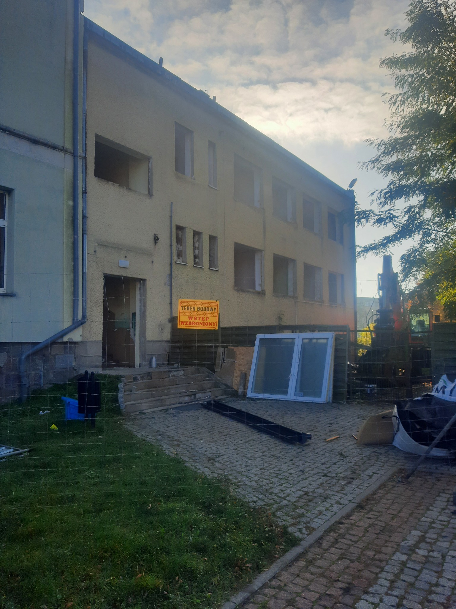 Budynek w trakcie prac rozbiórkowych, z wyjętymi oknami i widocznym znakiem ostrzegawczym 'Teren budowy, wstęp wzbroniony', oraz nowymi oknami przygotowanymi do montażu, widoczny sprzęt budowlany...