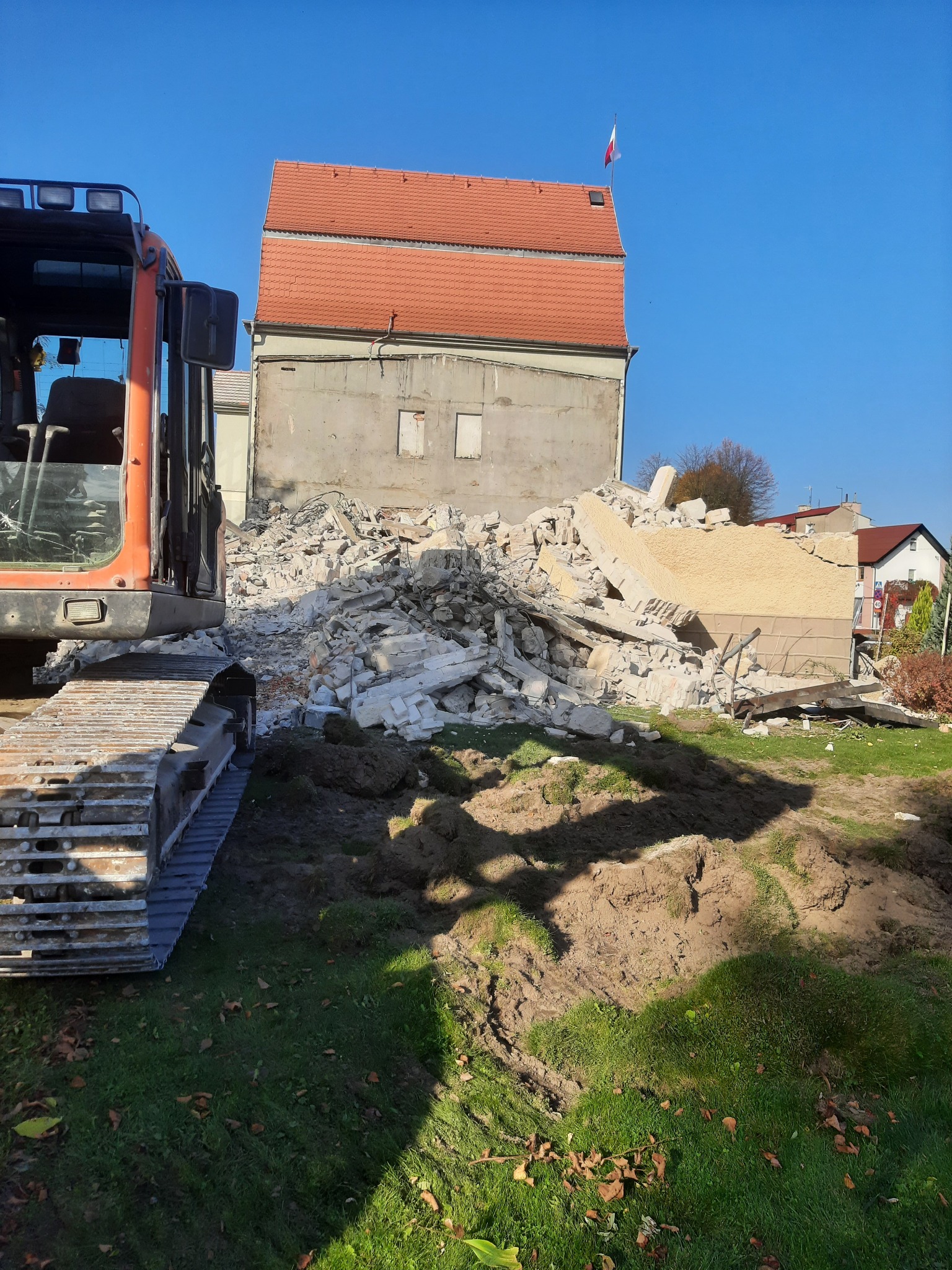 Pomarańczowa koparka gąsienicowa częściowo widoczna z lewej strony, na tle sterty gruzu po rozbiórce budynku, z widocznym fragmentem budynku z czerwoną dachówką i polską flagą na dachu, w słoneczny...