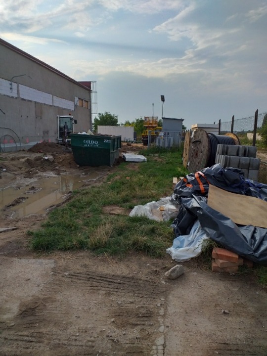 Teren budowy z kontenerem na odpady, mini koparką w tle, kablami w bębnach, kostką brukową i stertą gruzu przykrytą folią, widok na budynek przemysłowy i pochmurne niebo.