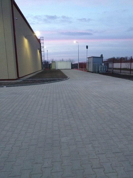 Ułożona kostka brukowa prowadząca do budynku przemysłowego z kontenerem i słupami oświetleniowymi w tle, widok w pochmurny dzień.