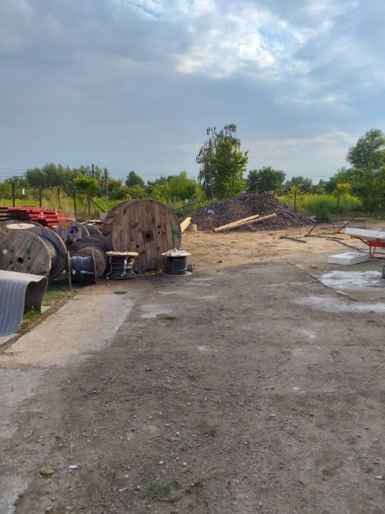 Składowisko materiałów budowlanych: drewniane szpule po kablach, sterta gruzu i ziemi z deskami, elementy konstrukcji stalowych w tle, pochmurne niebo.