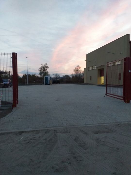 Teren po robotach ziemnych w Kliniskach Wielkich, z widocznymi śladami opon na piasku, ułożoną kostką brukową, ogrodzeniem z bramą, budynkiem w tle i zachodzącym słońcem.