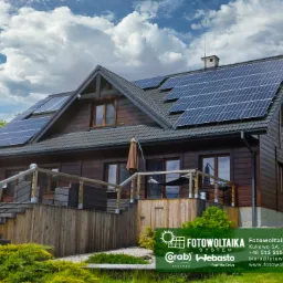 Dom z ciemnego drewna z panelami słonecznymi na dachu, widok z przodu, na pierwszym planie zieleń, błękitne niebo z chmurami w tle.