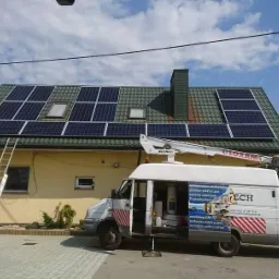 Montaż paneli słonecznych na dachu domu jednorodzinnego, widoczny samochód serwisowy z podnośnikiem i drabiny opierające się o ścianę budynku.