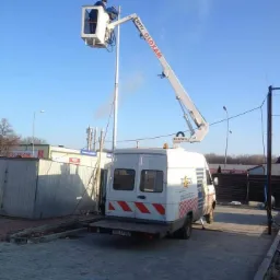 Pracownik na podnośniku koszowym przy słupie oświetleniowym, widoczny biały samochód z podnośnikiem na tle błękitnego nieba.