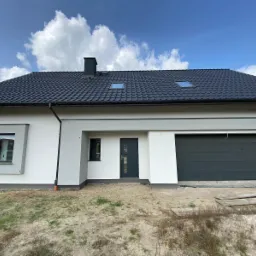 Nowoczesny dom jednorodzinny z grafitową bramą garażową segmentową, drzwiami zewnętrznymi w kolorze antracytu, oknami PCV z ciemnymi ramami i dachem z dachówki ceramicznej w odcieniach szarości...