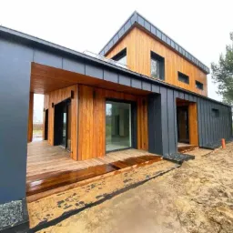 Nowoczesny dom z drewnianą elewacją, grafitowymi panelami i przesuwnymi drzwiami tarasowymi, widok z zewnątrz na werandę z drewnianym podestem.