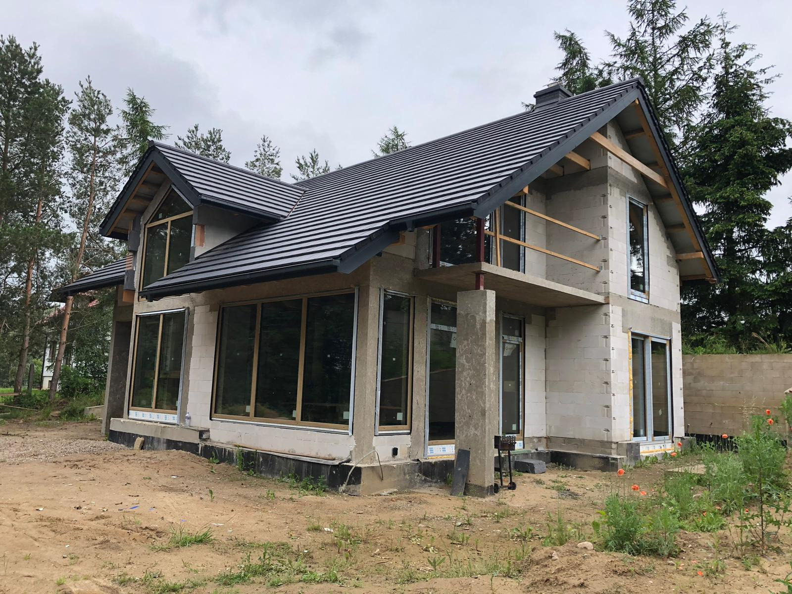 Surowy stan domu jednorodzinnego z zamontowanymi oknami z ramami w kolorze drewna, widoczne betonowe słupy podtrzymujące balkon i czarna dachówka na dachu. Na pierwszym planie ziemia i trawa.