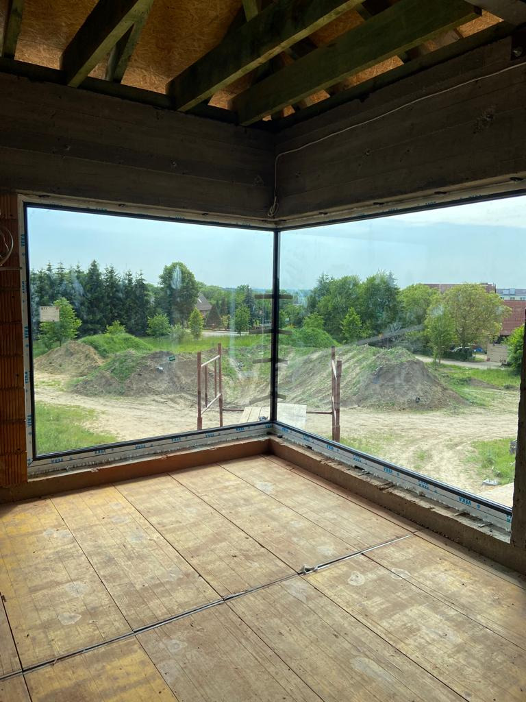 Narożne okno z widokiem na zielony krajobraz w surowym wnętrzu budynku w trakcie budowy, drewniana podłoga, widoczne elementy konstrukcyjne dachu.