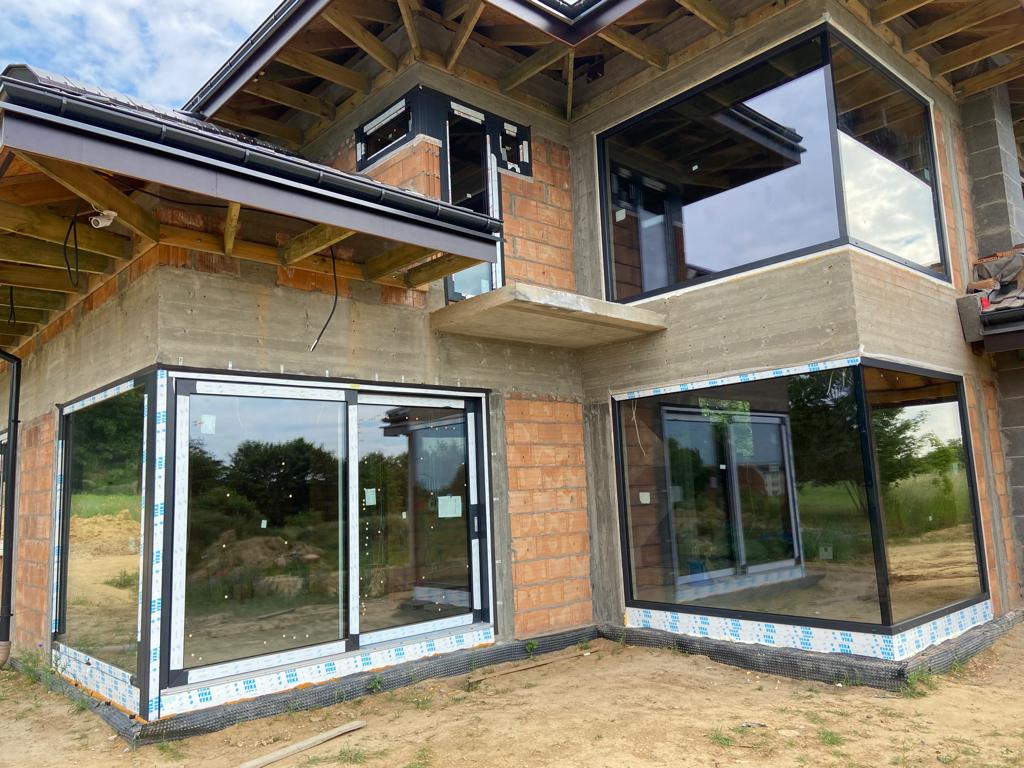 Surowy stan budynku z zamontowanymi oknami panoramicznymi z czarnymi ramami, widoczne cegły i beton, zabezpieczenie folią ochronną na profilach okiennych.