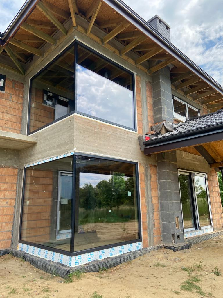 Narożne okno z czarną ramą w surowym stanie budynku, widoczne cegły i beton, odblask nieba na szybach.