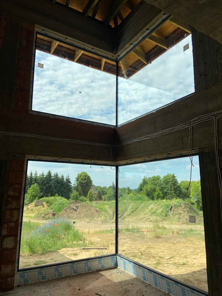 Narożne okno z czarną ramą w surowym stanie budynku, widok na zewnątrz z zielenią i niebem.