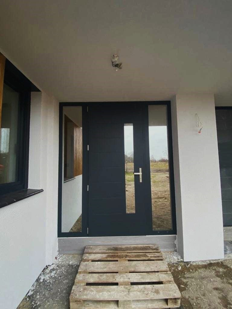 Nowoczesne, antracytowe drzwi wejściowe z przeszkleniem pionowym, zamontowane w nowym domu, widoczne z zewnątrz, z paletą drewnianą przed progiem.