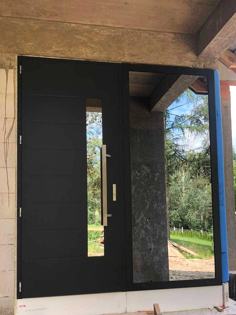 Nowoczesne, antracytowe drzwi wejściowe z bocznym przeszkleniem, zamontowane w surowym stanie budynku, widoczne elementy konstrukcyjne i zabezpieczenia.