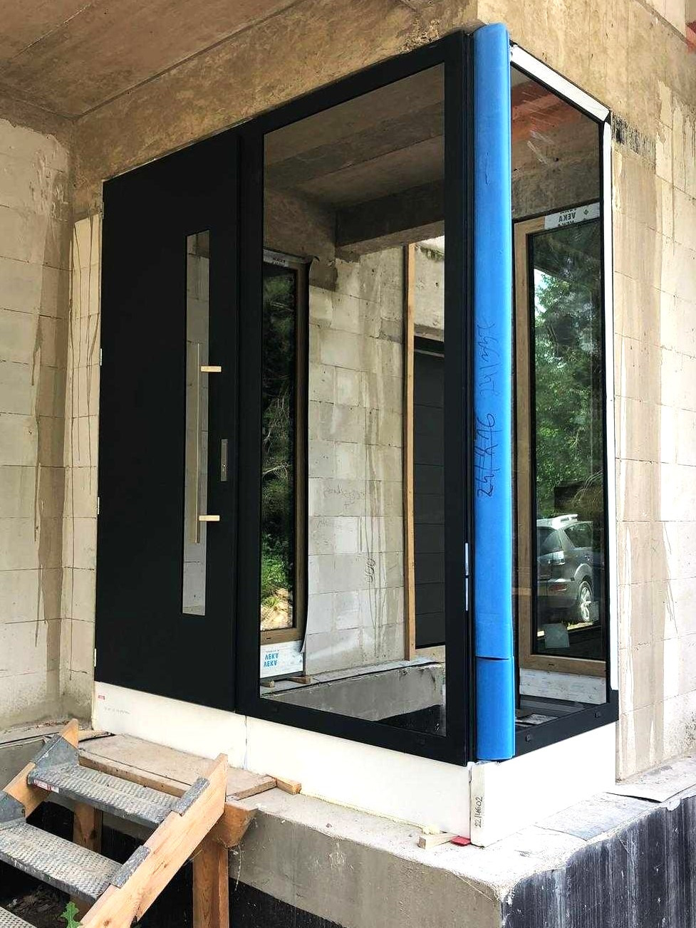 Czarne drzwi wejściowe z przeszkleniem, zamontowane w surowym stanie budynku, zabezpieczone niebieską folią ochronną, widoczne tymczasowe schodki.