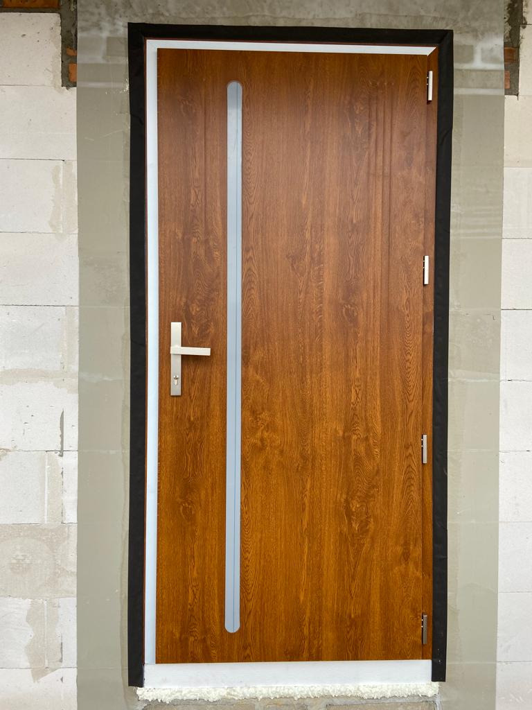 Brązowe drzwi wejściowe z pionowym przeszkleniem, zamontowane w surowym murze z betonu komórkowego, widoczne czarne uszczelki wokół ościeżnicy.