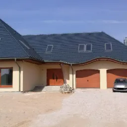 Nowoczesny dom jednorodzinny z ciemnym dachem z licznymi oknami dachowymi, jasną elewacją i dwustanowiskowym garażem z brązowymi bramami, na podjeździe zaparkowany srebrny samochód.