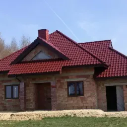 Dom jednorodzinny w trakcie budowy z nowym, czerwonym pokryciem dachowym z dachówki ceramicznej i ceglanym kominem, widoczne rynny i częściowo wykończona elewacja z cegły.