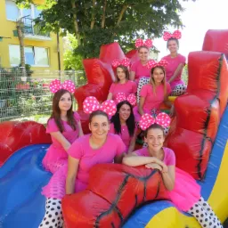 Grupa animatorek w różowych strojach i opaskach z kokardami w kropki, siedzących na dmuchanym zamku z motywem uśmiechniętej twarzy, na tle budynku i drzew.