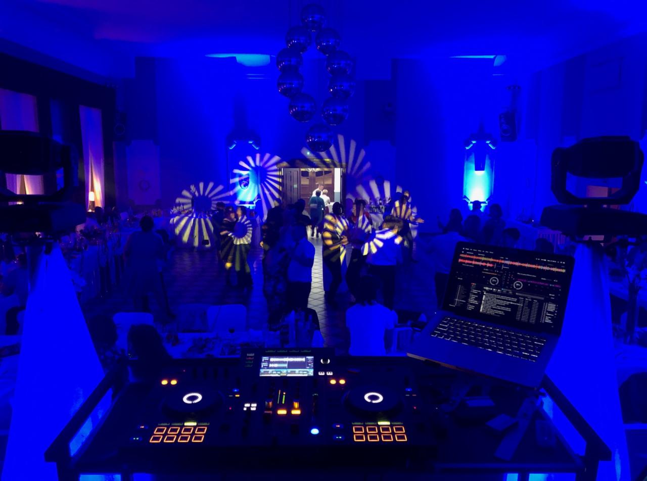 DJ na weselu, widok zza konsoli DJ-skiej z laptopem, w tle tańczący goście oświetleni niebieskim światłem i ruchomymi reflektorami z wzorem promieni, nad nimi kule dyskotekowe.