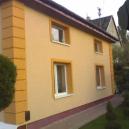 Dwukondygnacyjny dom z elewacją w odcieniach beżu i żółci, z dekoracyjnymi narożnikami w kolorze żółtym, widoczny z boku w otoczeniu zieleni.