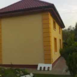 Żółta elewacja domu jednorodzinnego z brązowym dachem, ozdobnymi panelami narożnymi i niskim, białym cokołem, widoczna z perspektywy ogrodu.