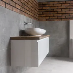 Nowoczesna łazienka z umywalką nablatową, białą szafką pod umywalką z drewnianym blatem i ścianą wykończoną szarą płytką oraz cegłą.