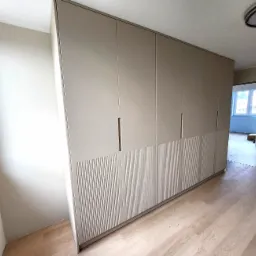 Beżowa szafa wnękowa z pionowymi żłobieniami na dolnych panelach, zamontowana w pokoju z drewnianą podłogą.