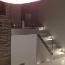 Nowoczesne schody z białymi stopniami, podświetleniem LED i szklaną balustradą, widoczne z dołu, obok ściany z cegły dekoracyjnej.