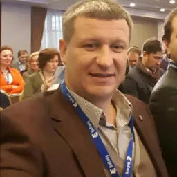 Mężczyzna w brązowej marynarce i identyfikatorem na niebieskiej smyczy z logo firmy, uśmiecha się do kamery na tle sali konferencyjnej z innymi uczestnikami.