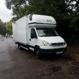 Biały samochód dostawczy Iveco z zabudową kontenerową stoi na mokrej ulicy w pochmurny dzień, widoczne drzewa w tle.