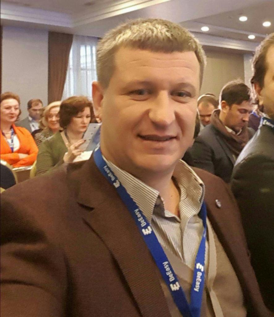 Mężczyzna w brązowej marynarce i identyfikatorem na niebieskiej smyczy z logo firmy, uśmiecha się do kamery na tle sali konferencyjnej z innymi uczestnikami.