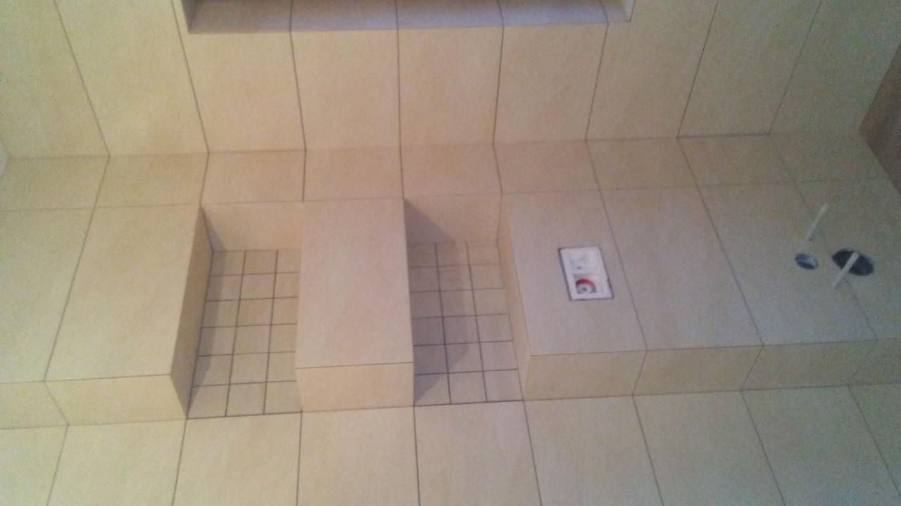 Wykończona łazienka z beżowymi płytkami, wnęki prysznicowe z ciemniejszymi płytkami na dnie, widoczne otwory na instalacje.