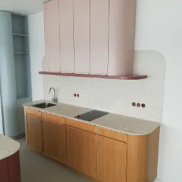 Nowoczesna kuchnia z drewnianymi szafkami dolnymi o zaokrąglonych krawędziach, jasnoróżowymi szafkami górnymi, blatem z lastryko i minimalistyczną armaturą, uzupełniona o półkę w kolorze bordo.