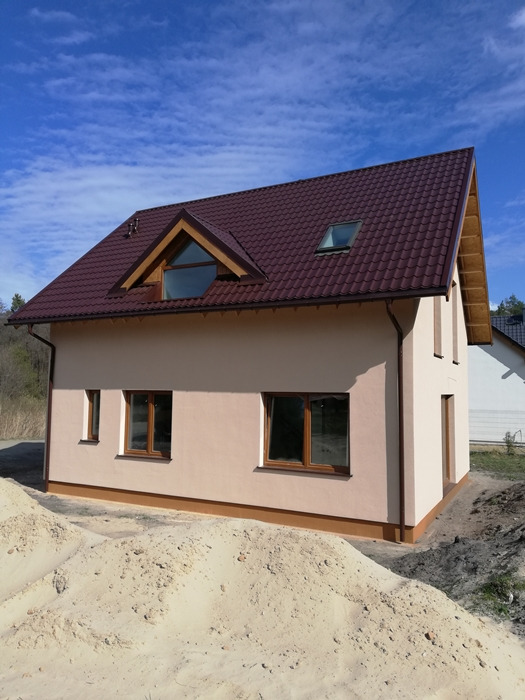 Niedawno wybudowany dom z brązowym dachem z blachodachówki, jasnobeżową elewacją i brązowymi oknami, widoczny stos piasku przed budynkiem.