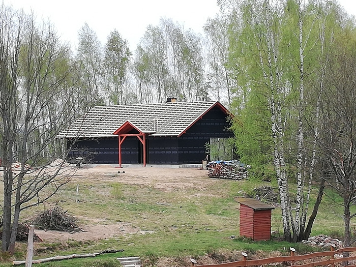 Budowa domu w lesie: czarna elewacja, dach w trakcie układania, czerwony podcień wejściowy, sterta drewna opałowego w tle.
